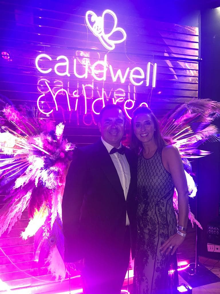 Smiling couple in formal wear standing before a vibrant purple Caudwell Children neon sign.