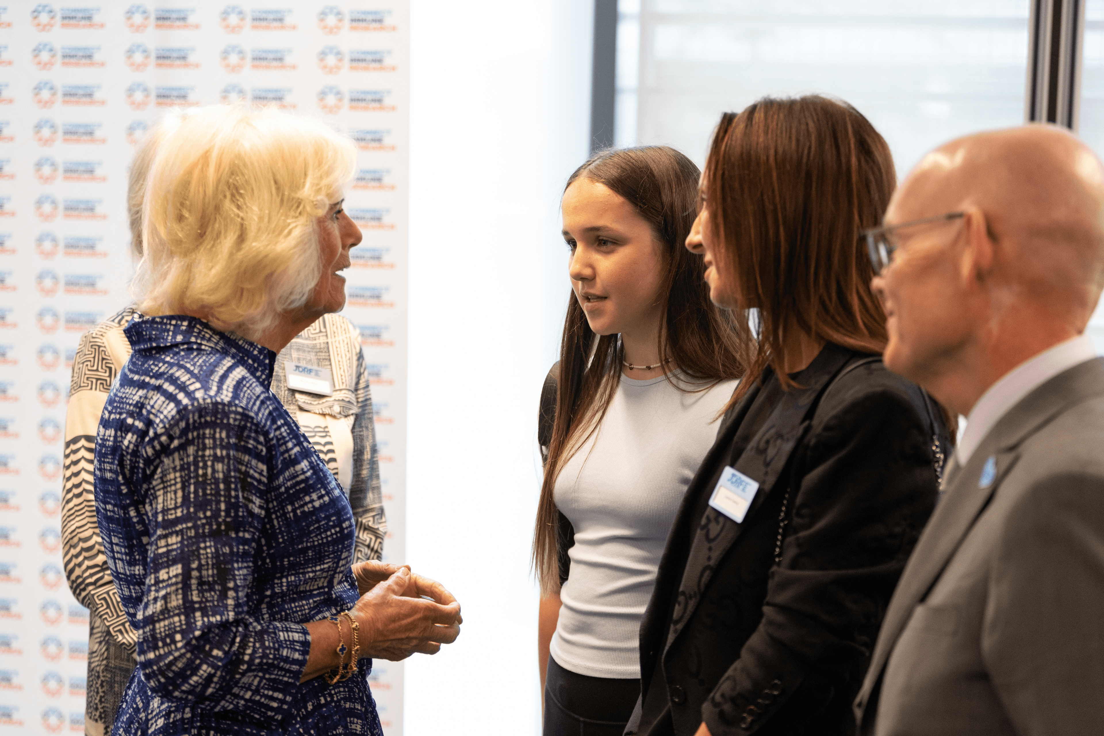 Queen Camilla in a blue patterned dress speaks with a young girl and two adults.