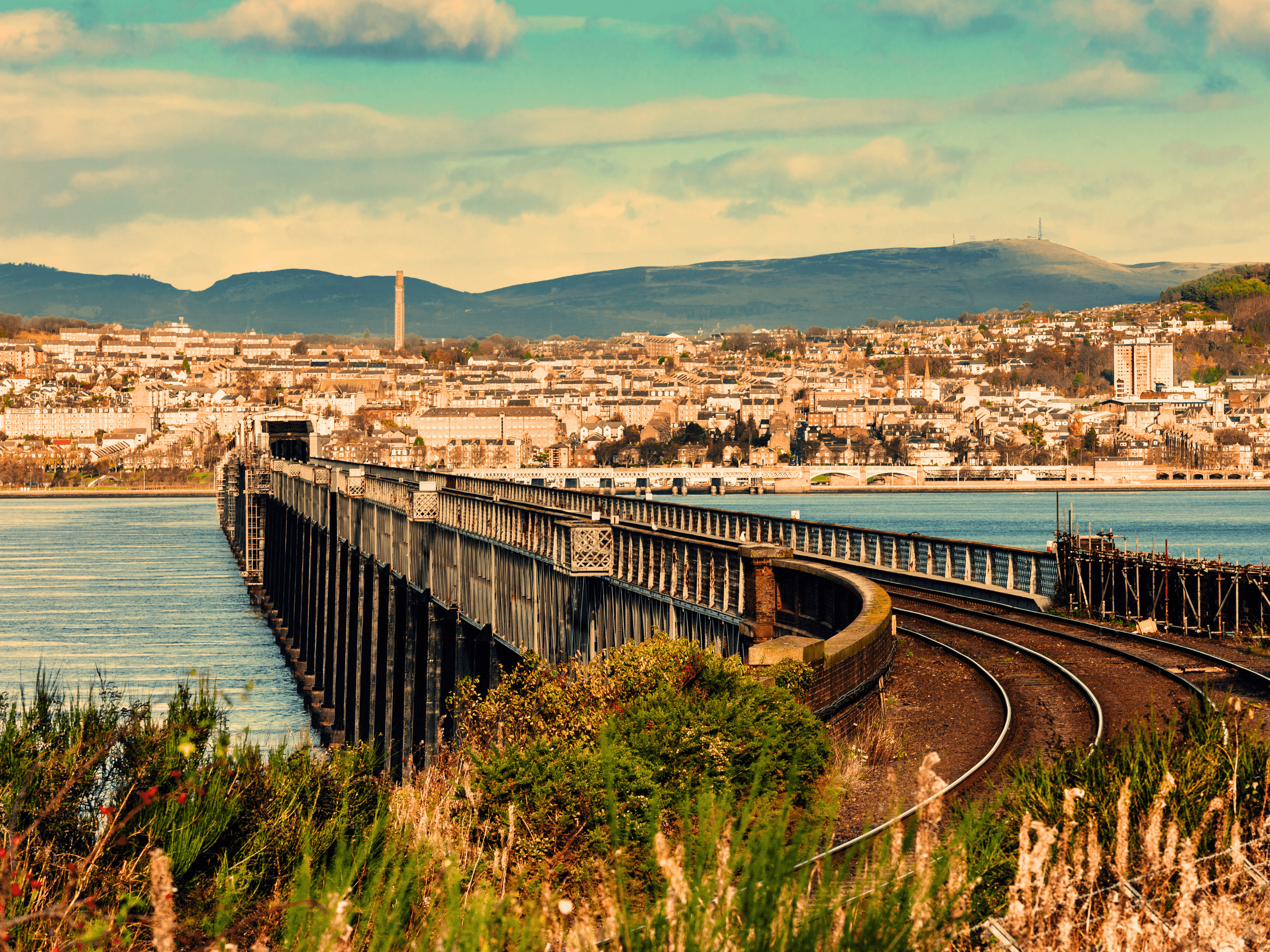 Curved railway tracks lead onto a long bridge spanning water toward a city and hills.