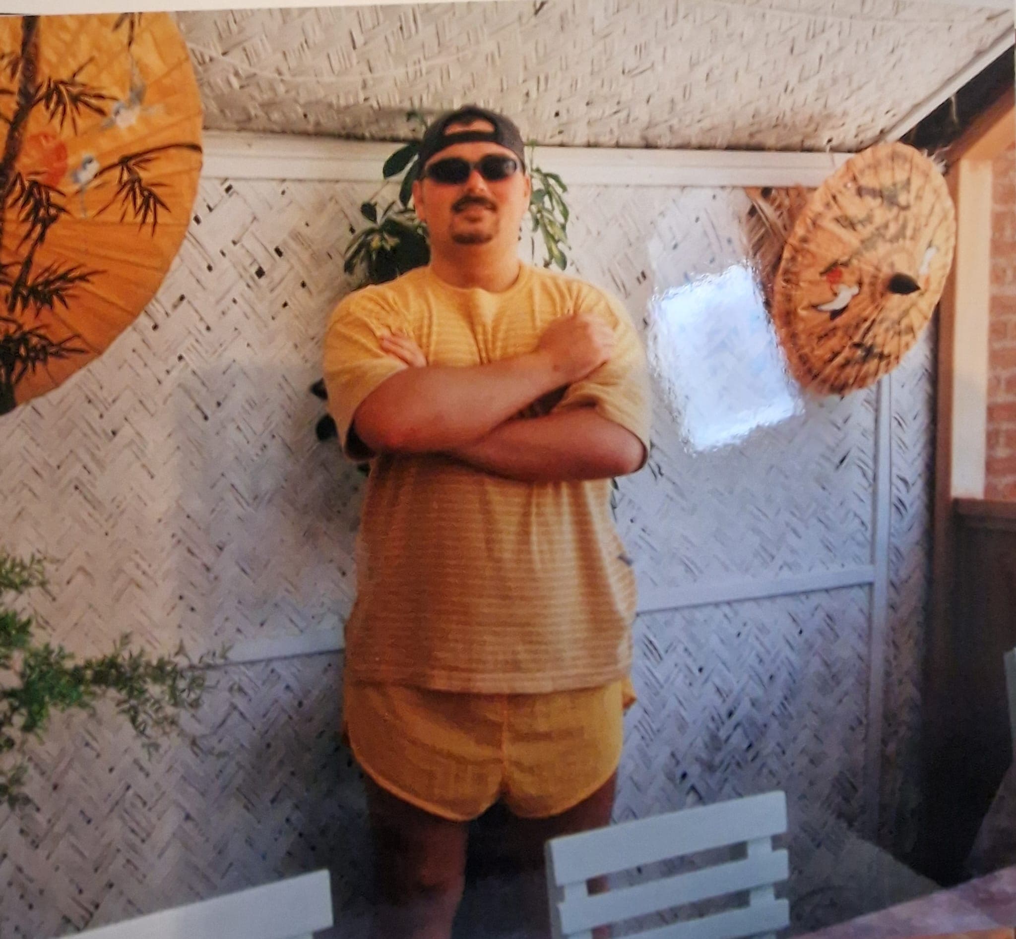 Man in yellow outfit, sunglasses, and backward cap poses with arms crossed against woven matting.